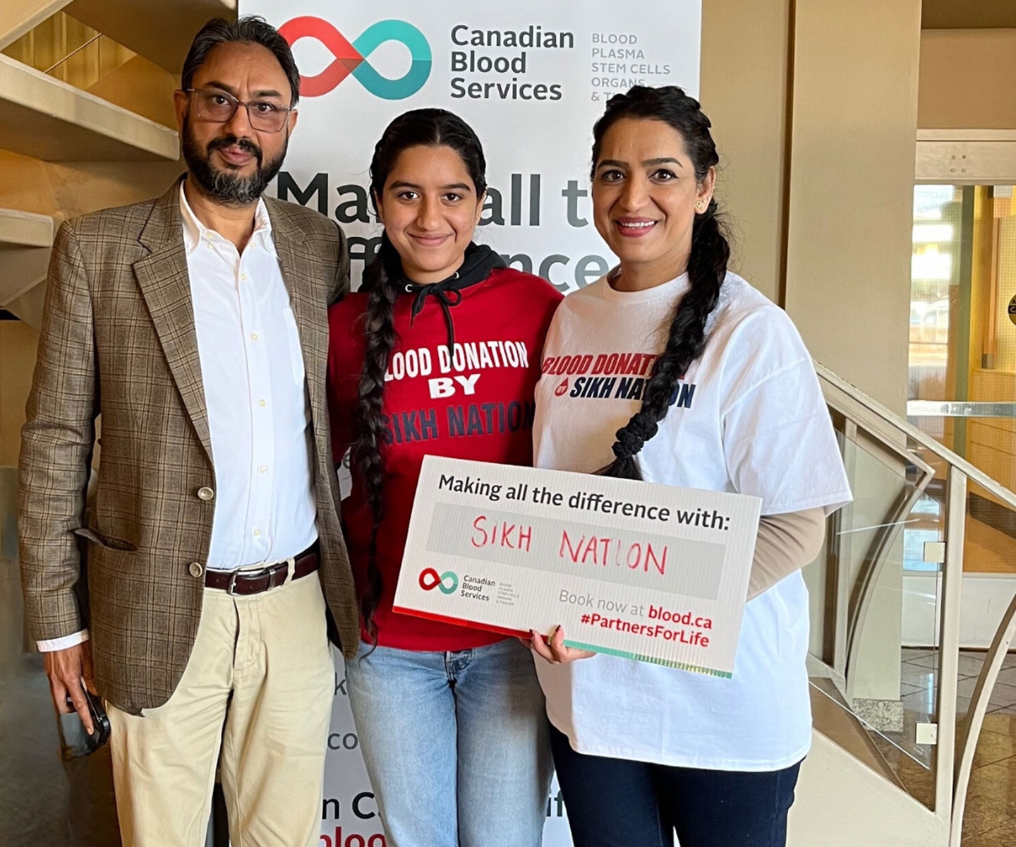 Une famille de trois personnes se tient ensemble derrière une pancarte sur laquelle est inscrit « Making all the difference with: Sikh Nation ». Deux d’entre elles portent un t-shirt Blood donation by Sikh Nation.