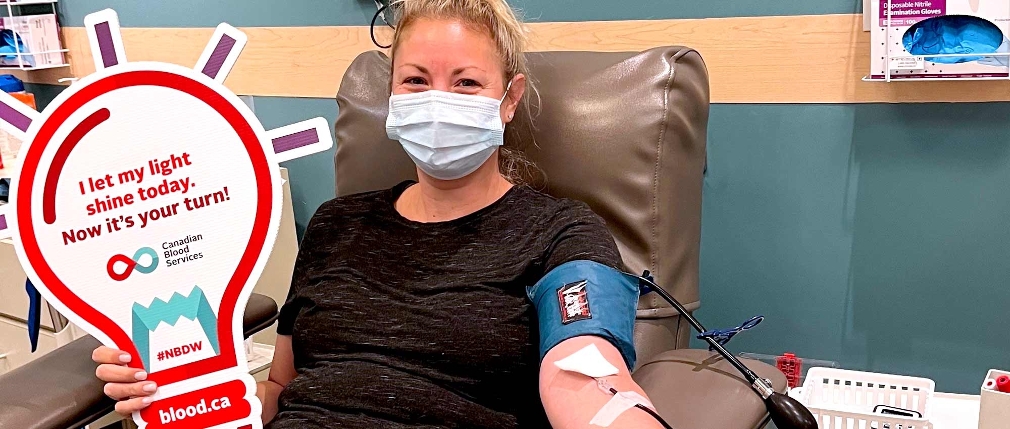 Blood donor in chair with sign shaped like lightbulb that says “I let my light shine today. Now it’s your turn.”