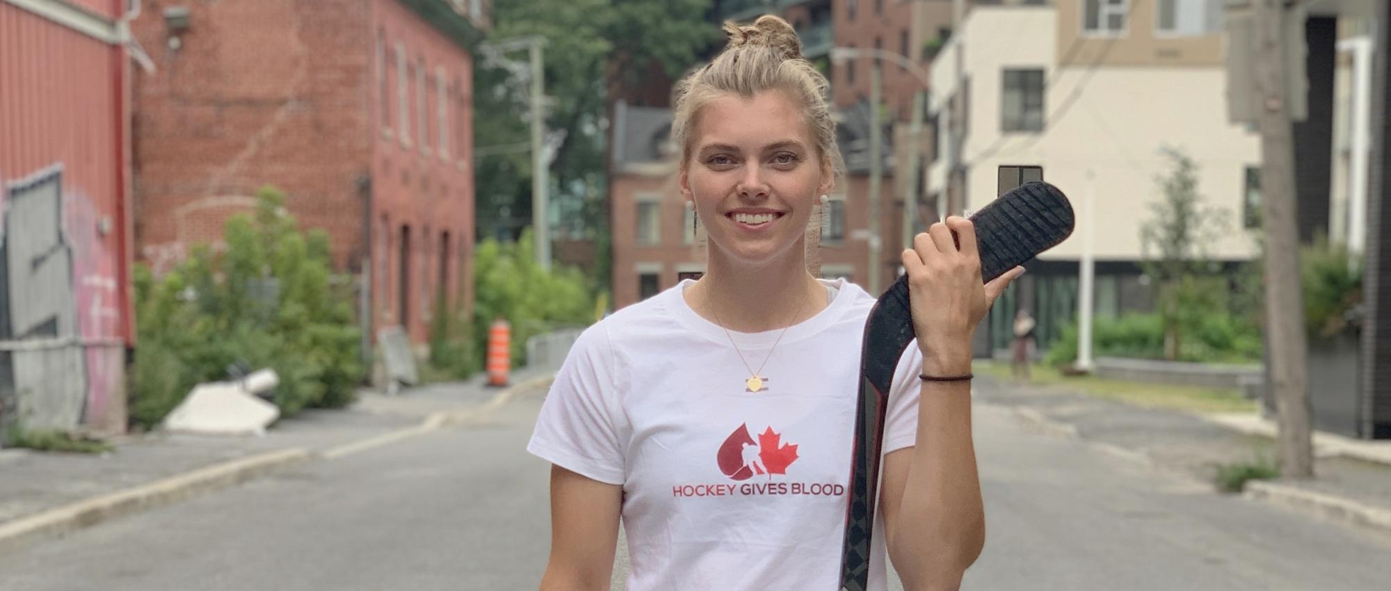 Image of a white blonde girl wearing a Hockey Gives Blood t-shirt and holding a hockey stick.