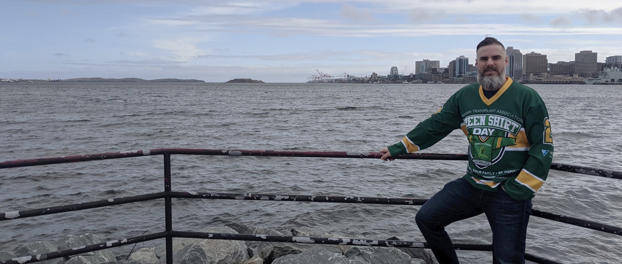 A man wearing a green shirt day jersey, in front of a body of water with a city skyline in the background.