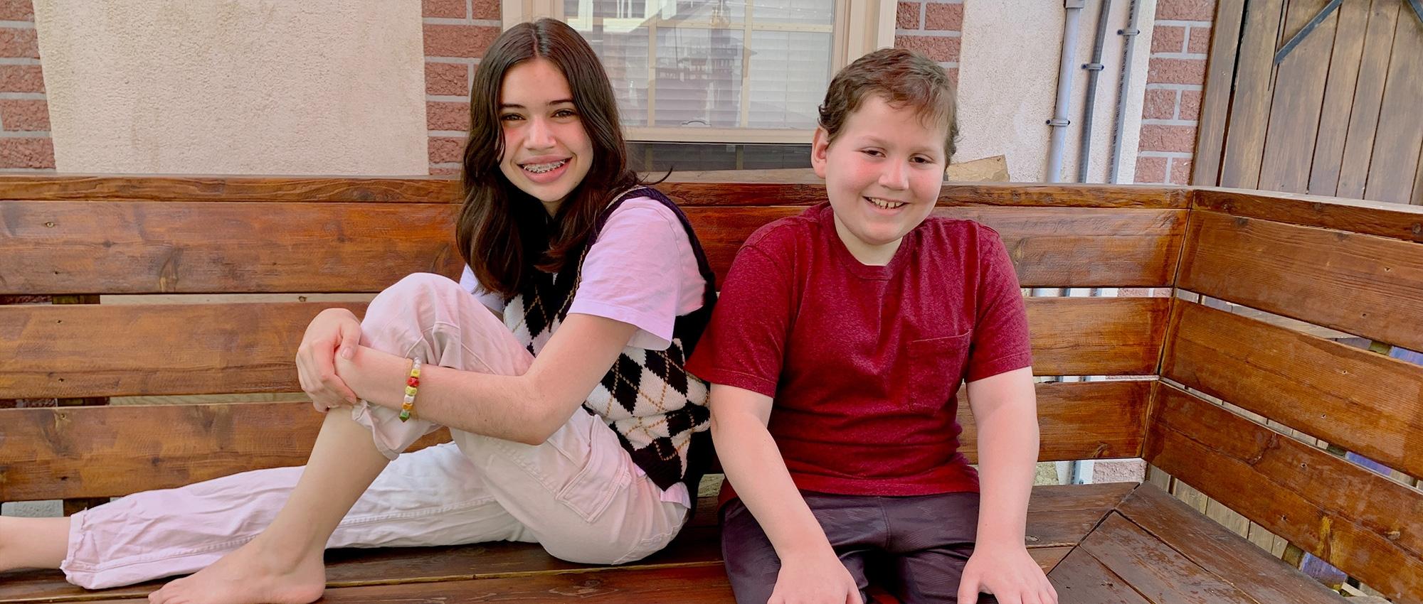 A sister and brother sit together on a bench outside their home.