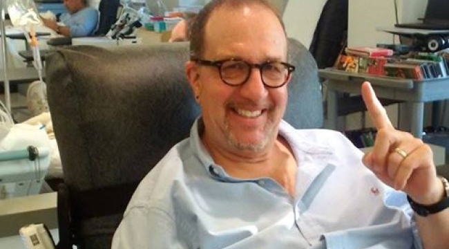 Image of Larry sitting in a donation chair donating blood holding up a one finger