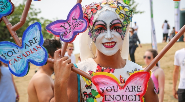 A man in white makeup decorated with colourful butterflies at a beach during a Pride celebration.