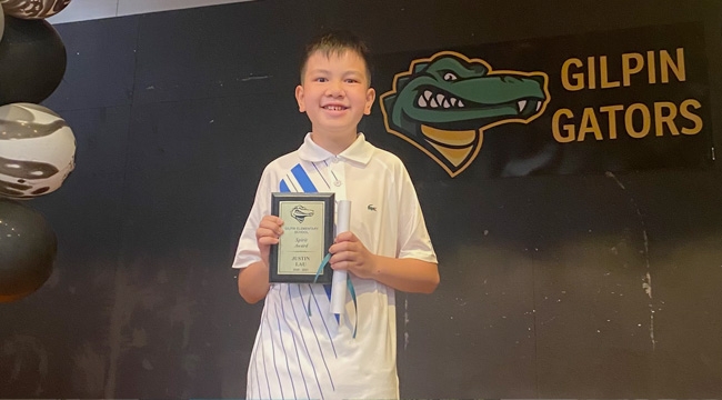 An Asian boy smiles as he holds his award plaque in front of a black background with “Gilpin Gators” inscription and logo. 